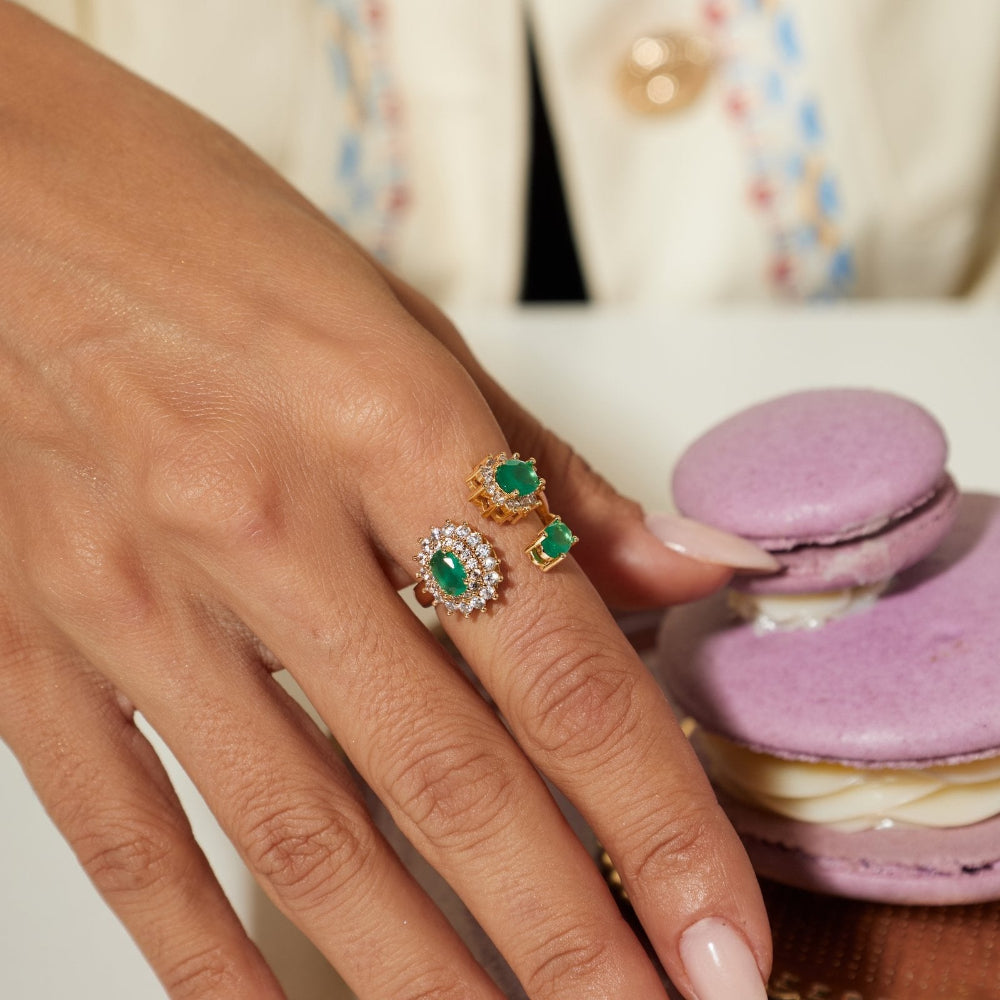 A close-up of a hand wearing two gold rings with green emerald-cut stones and small clear accent stones, posed over a purple macaron on a white surface.