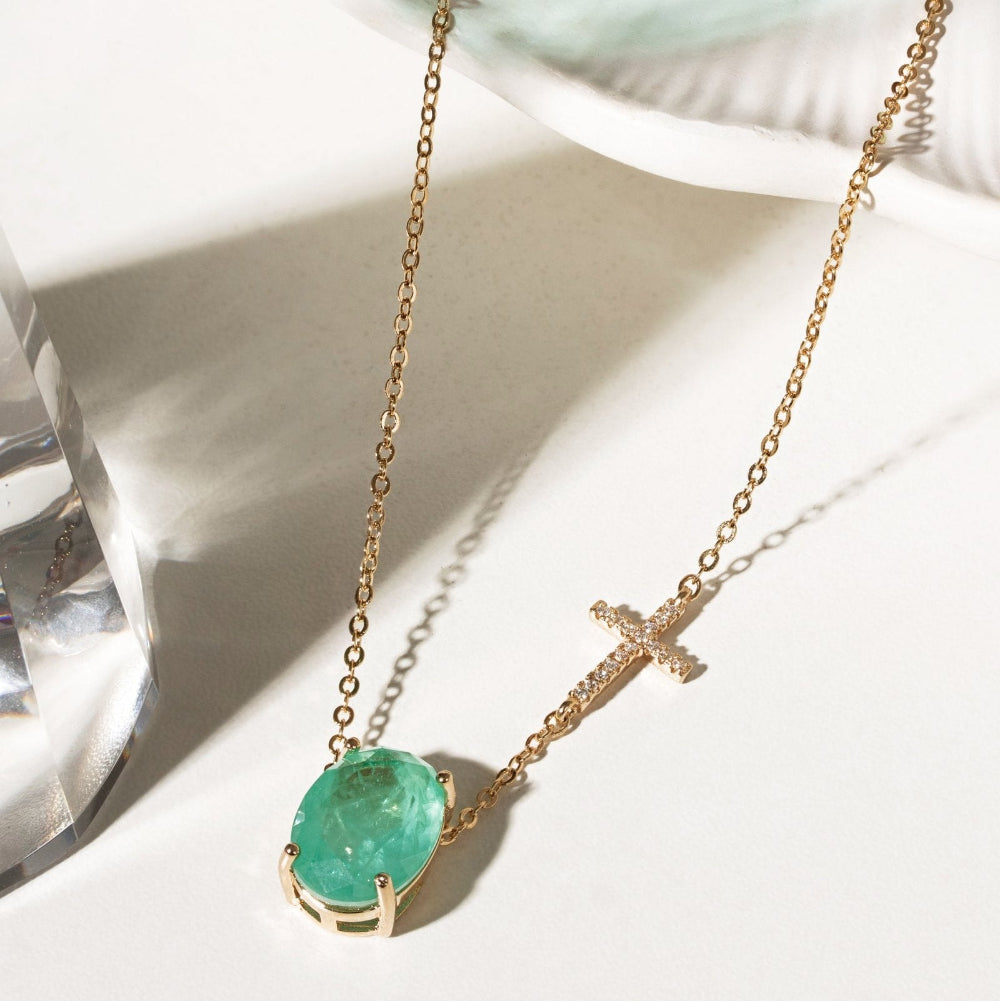 A gold chain necklace featuring a green oval gemstone pendant and a small gold cross charm, displayed on a white surface with soft shadows.