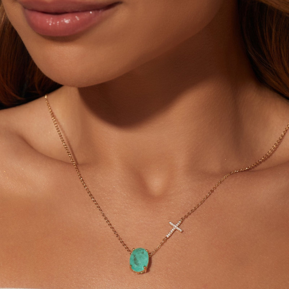 A close-up of a model wearing a delicate rose gold necklace with a small silver cross charm and a turquoise oval pendant.