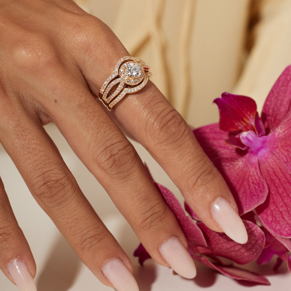 A close-up lifestyle shot of a woman's hand wearing a rose gold ring with a clear round-cut center stone and pavé accents, styled with a pink orchid flower against a soft beige blouse background.