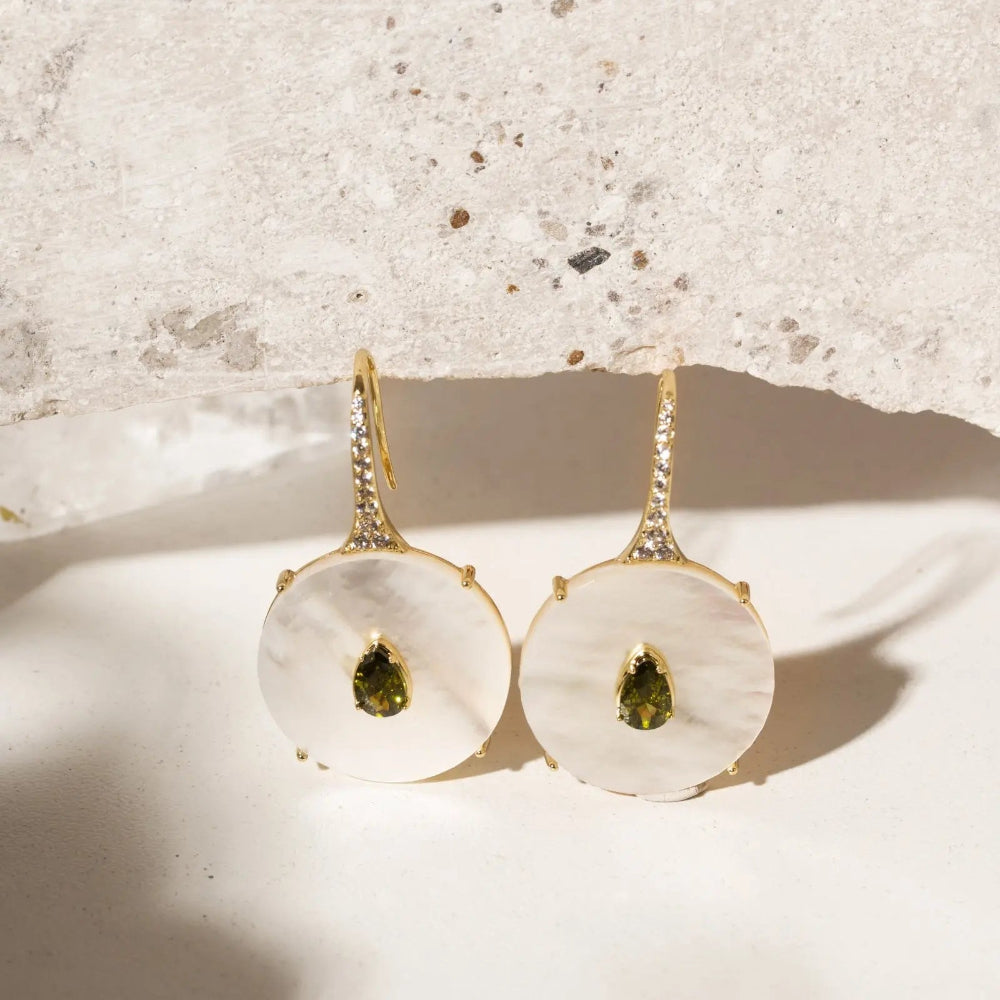 A pair of gold drop earrings with large white mother-of-pearl disc pendants, each featuring a small green teardrop gemstone in the center, displayed on a light surface with a textured stone slab behind them.