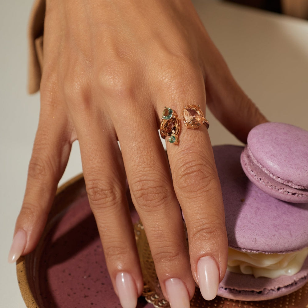 A close-up of a hand wearing a rose gold ring with green and clear stones, resting near a purple macaron on a speckled pink plate.