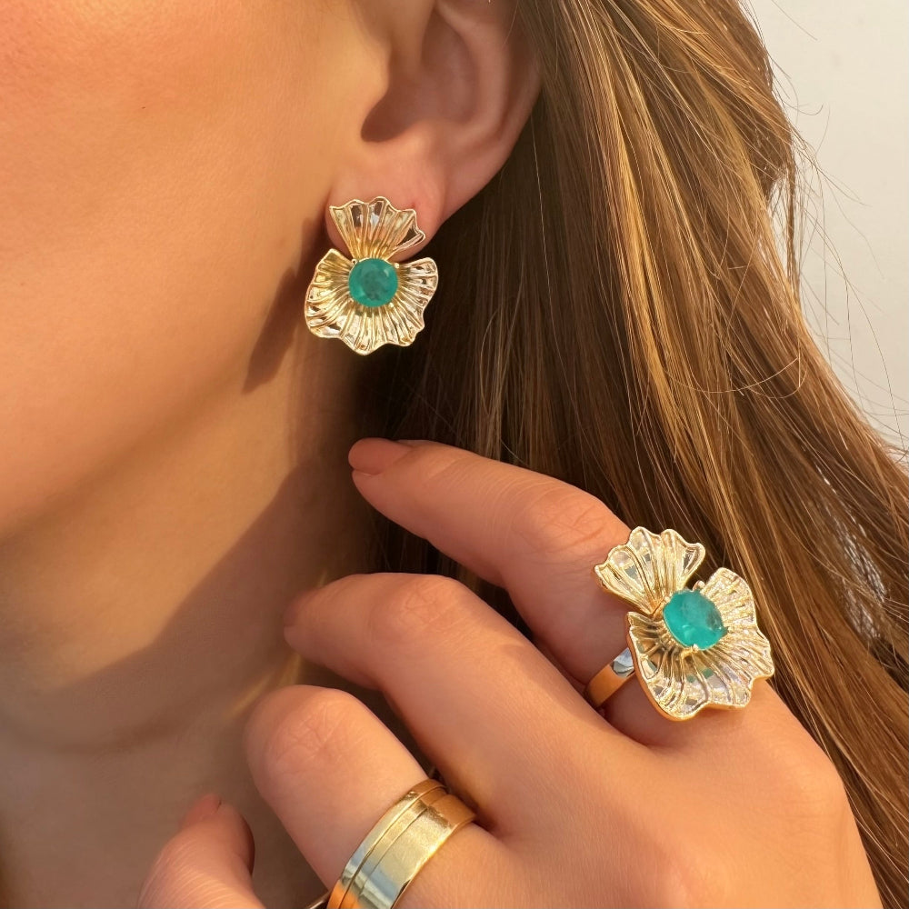 A close-up lifestyle shot of a model wearing matching gold flower-shaped earrings and rings, each featuring a round turquoise-colored center stone.